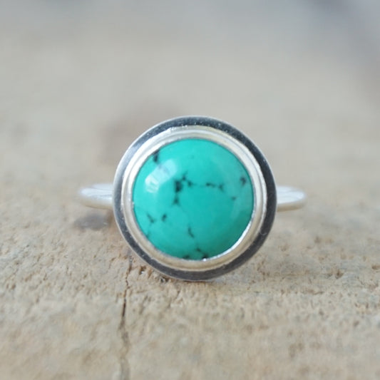 A sterling silver ring featuring a round turquoise cabochon stone with natural dark veining, set against a neutral, textured beige background.