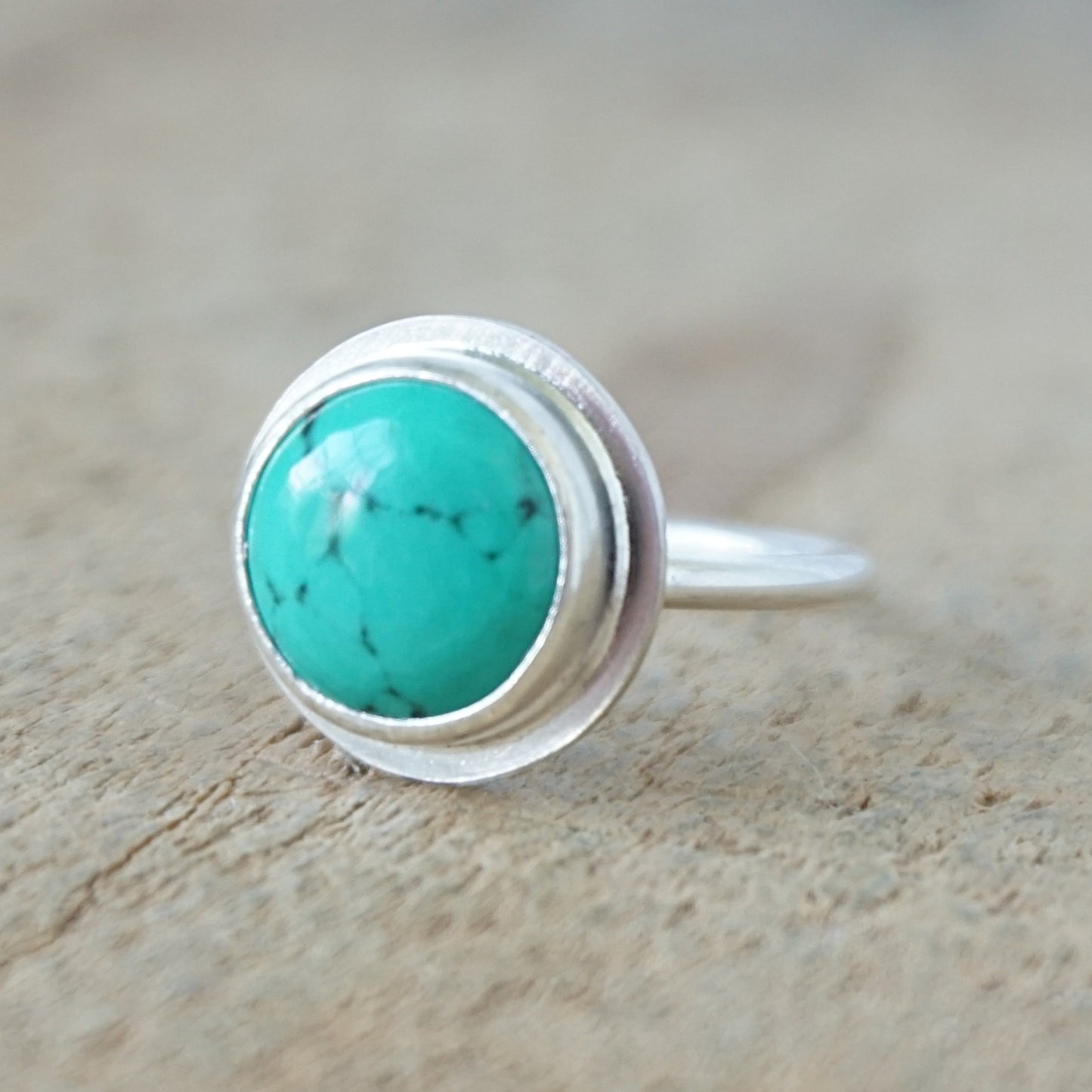 A sterling silver ring featuring a round turquoise cabochon stone with natural dark veining, set against a neutral, textured beige background.