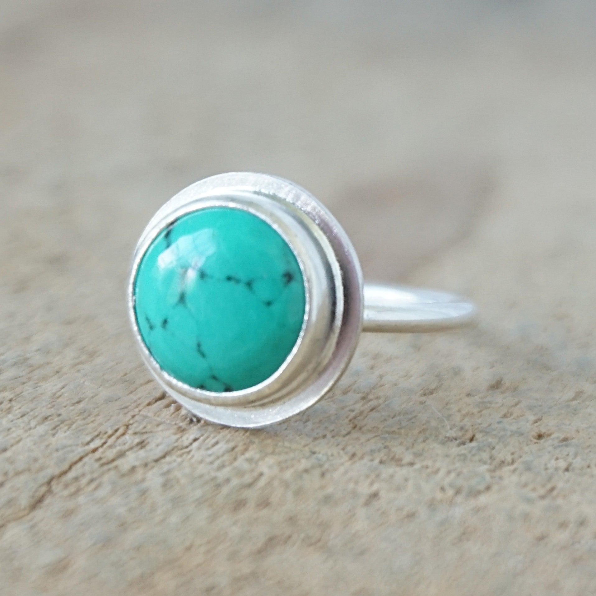 A sterling silver ring featuring a round turquoise cabochon stone with natural dark veining, set against a neutral, textured beige background.