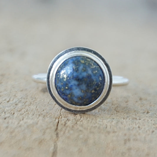 A sterling silver ring featuring a round bezel-set blue lapis lazuli stone with visible golden pyrite inclusions, resting on a textured beige surface.