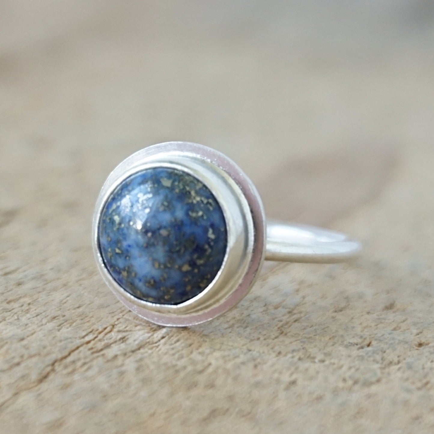 A sterling silver ring featuring a round cabochon of blue lapis lazuli with visible golden pyrite flecks, set against a neutral, textured beige background.