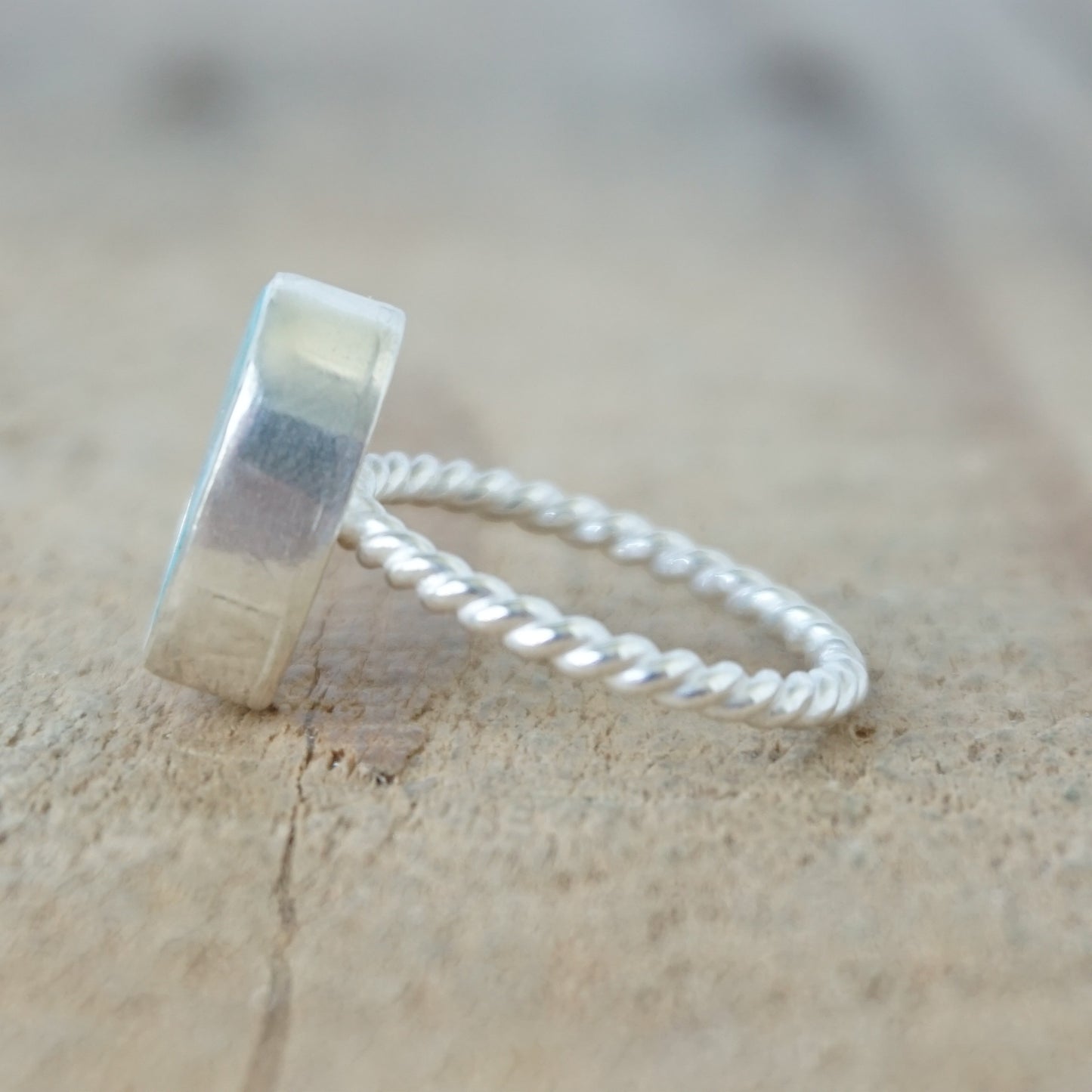 side view of A silver ring featuring an oval turquoise stone with natural matrix inclusions, set on a twisted rope-style silver band, on a wooden surface.