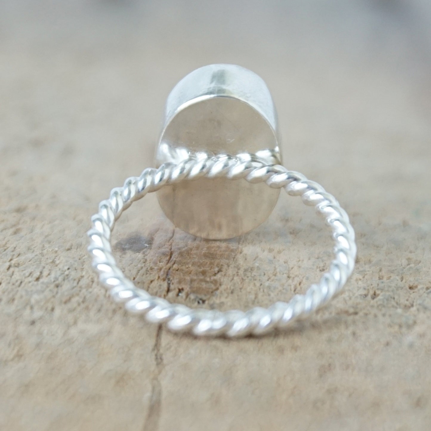 back view of A silver ring featuring an oval turquoise stone with natural matrix inclusions, set on a twisted rope-style silver band, on a wooden surface.