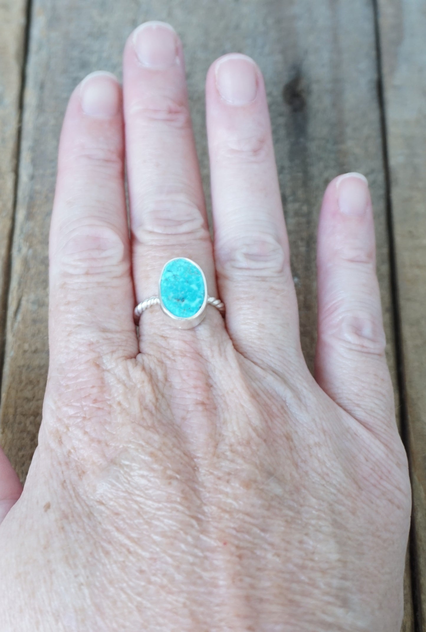 A silver ring with an oval turquoise cabochon stone worn on a person's ring finger, shown against a rustic wooden background.