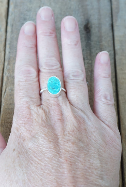 A silver ring with an oval turquoise cabochon stone worn on a person's ring finger, shown against a rustic wooden background.