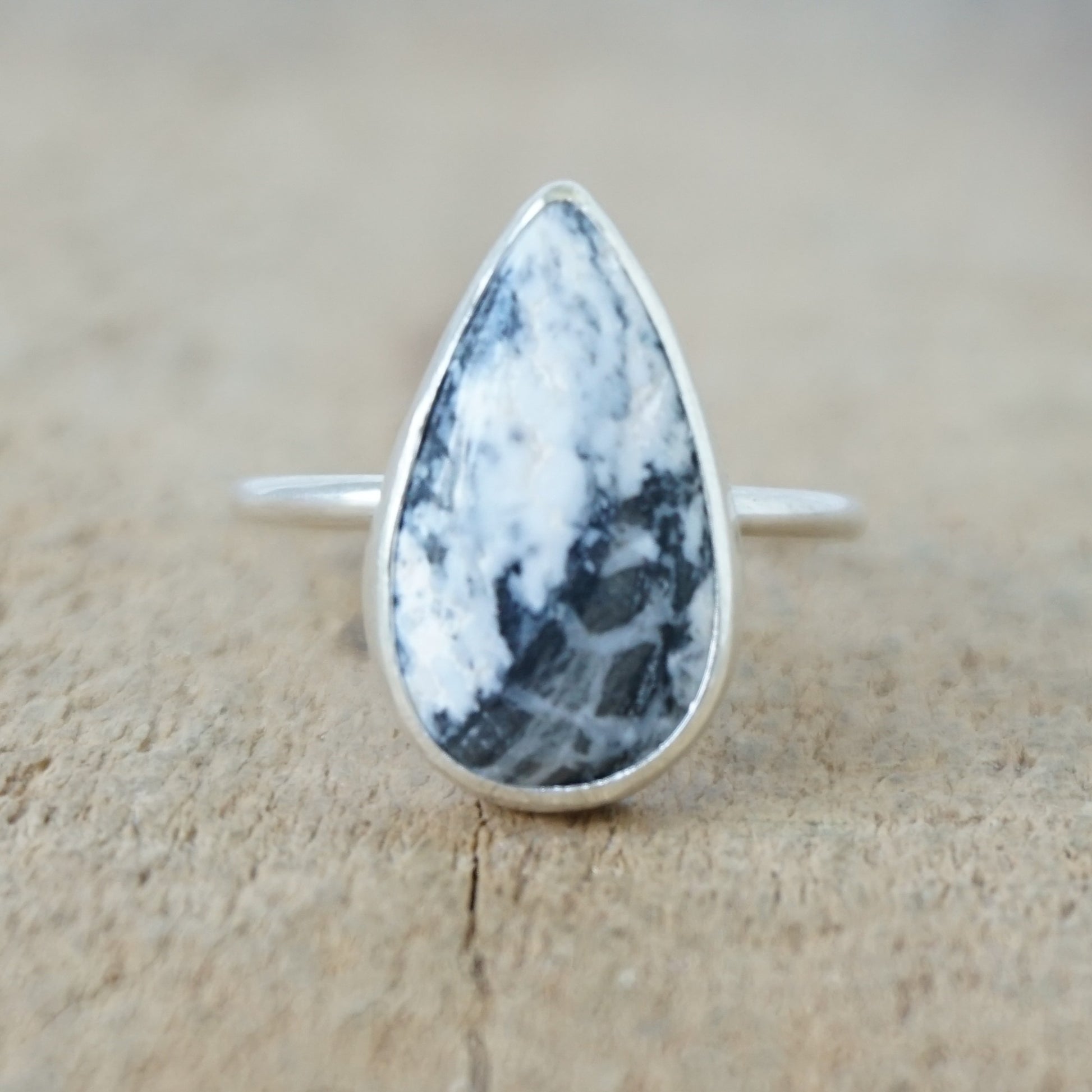 A silver ring featuring a teardrop-shaped gemstone with a white and black marbled pattern, on a wooden surface.