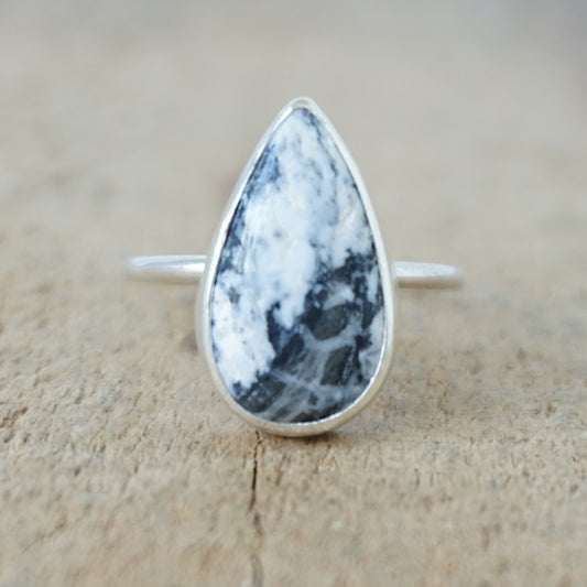 A silver ring featuring a teardrop-shaped gemstone with a white and black marbled pattern, on a wooden surface.