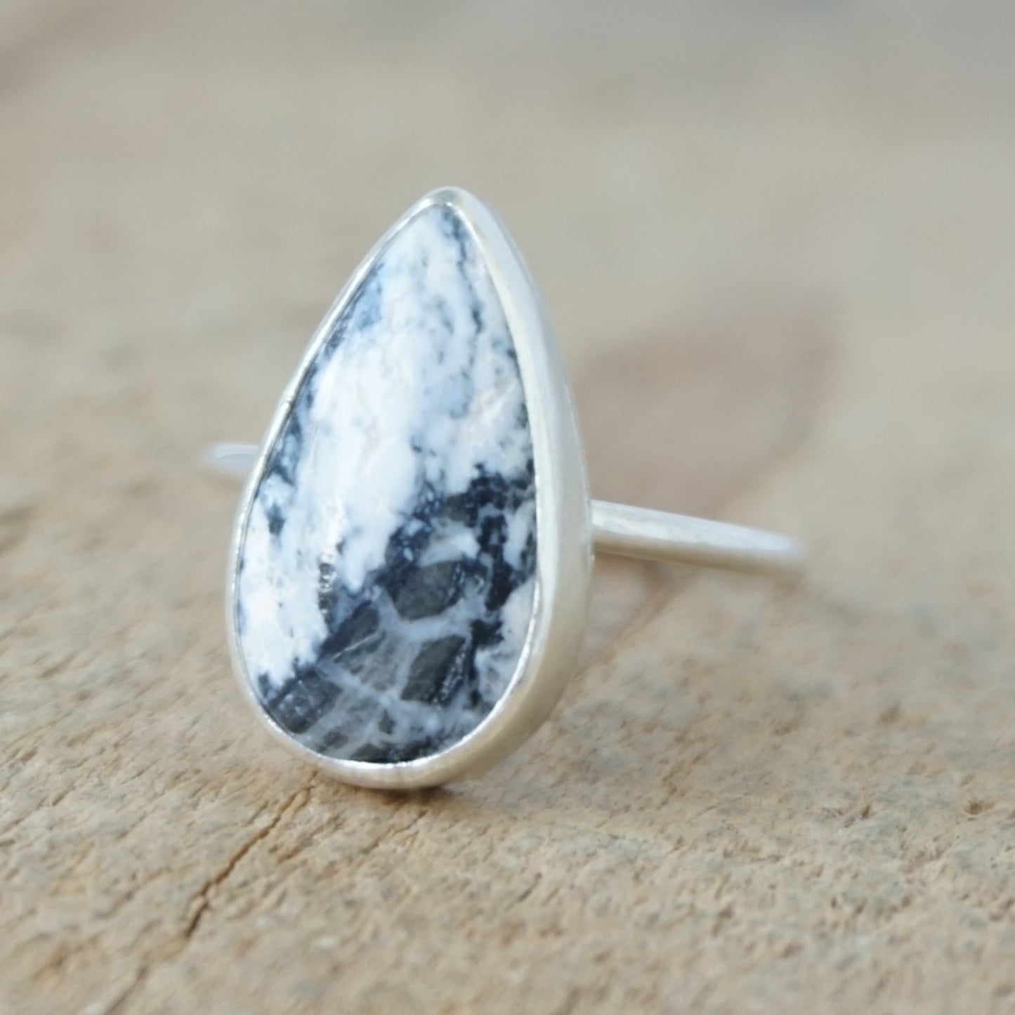 A silver ring featuring a teardrop-shaped gemstone with a white and black marbled pattern, on a wooden surface.