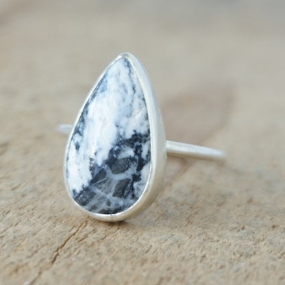 A silver ring featuring a teardrop-shaped gemstone with a white and black marbled pattern, on a wooden surface.