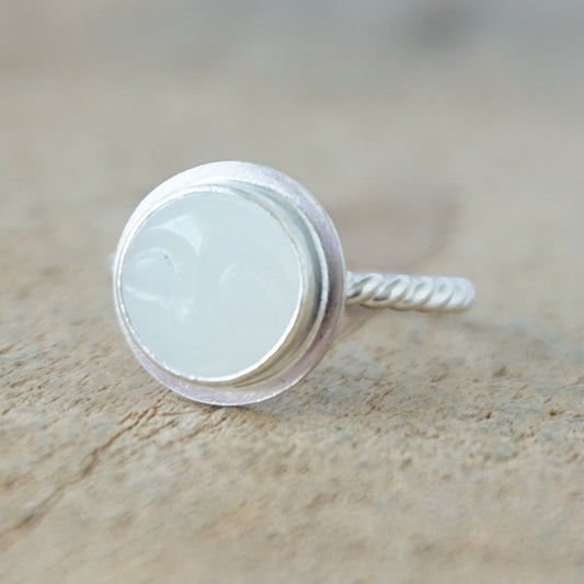 A sterling silver ring featuring a round bezel-set white stone with a carved face, on a wooden surface.
