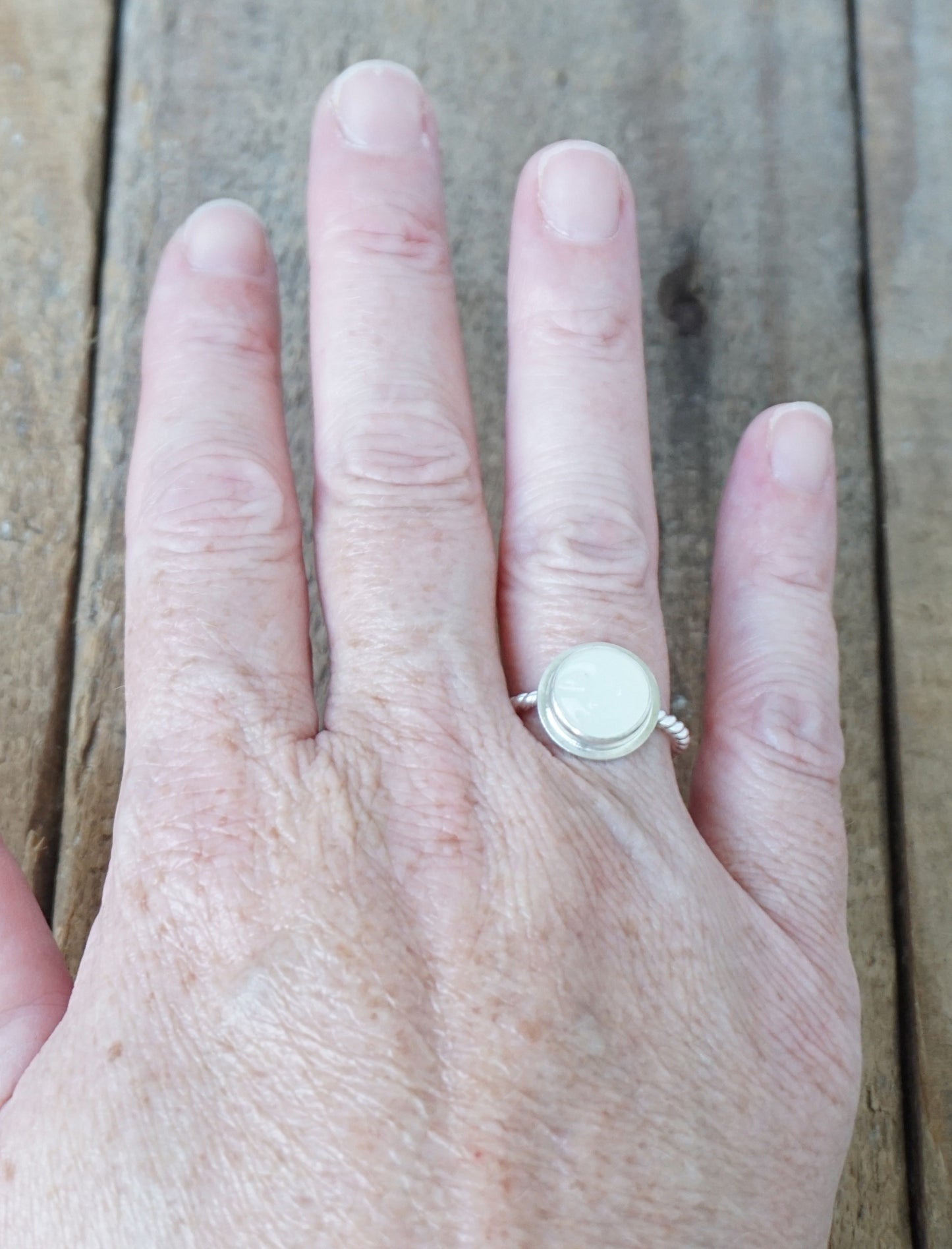 hand wearing A sterling silver ring featuring a round bezel-set white stone with a carved face, on a wooden surface.