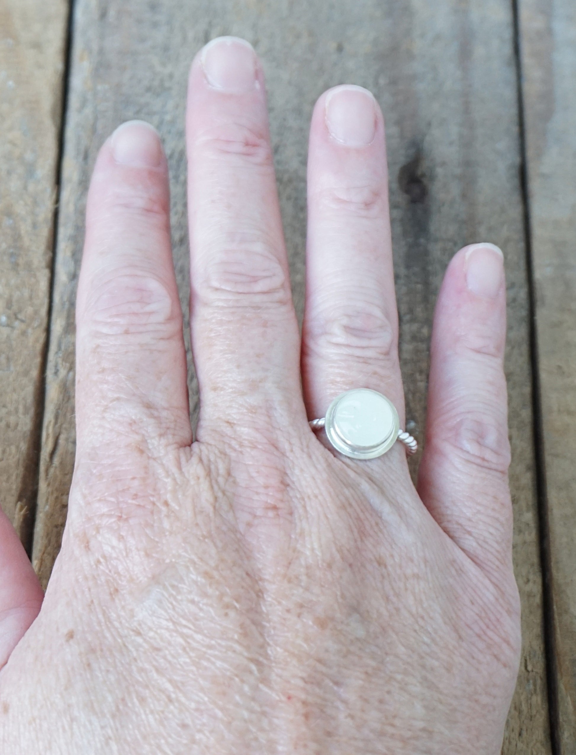 hand wearing A sterling silver ring featuring a round bezel-set white stone with a carved face, on a wooden surface.