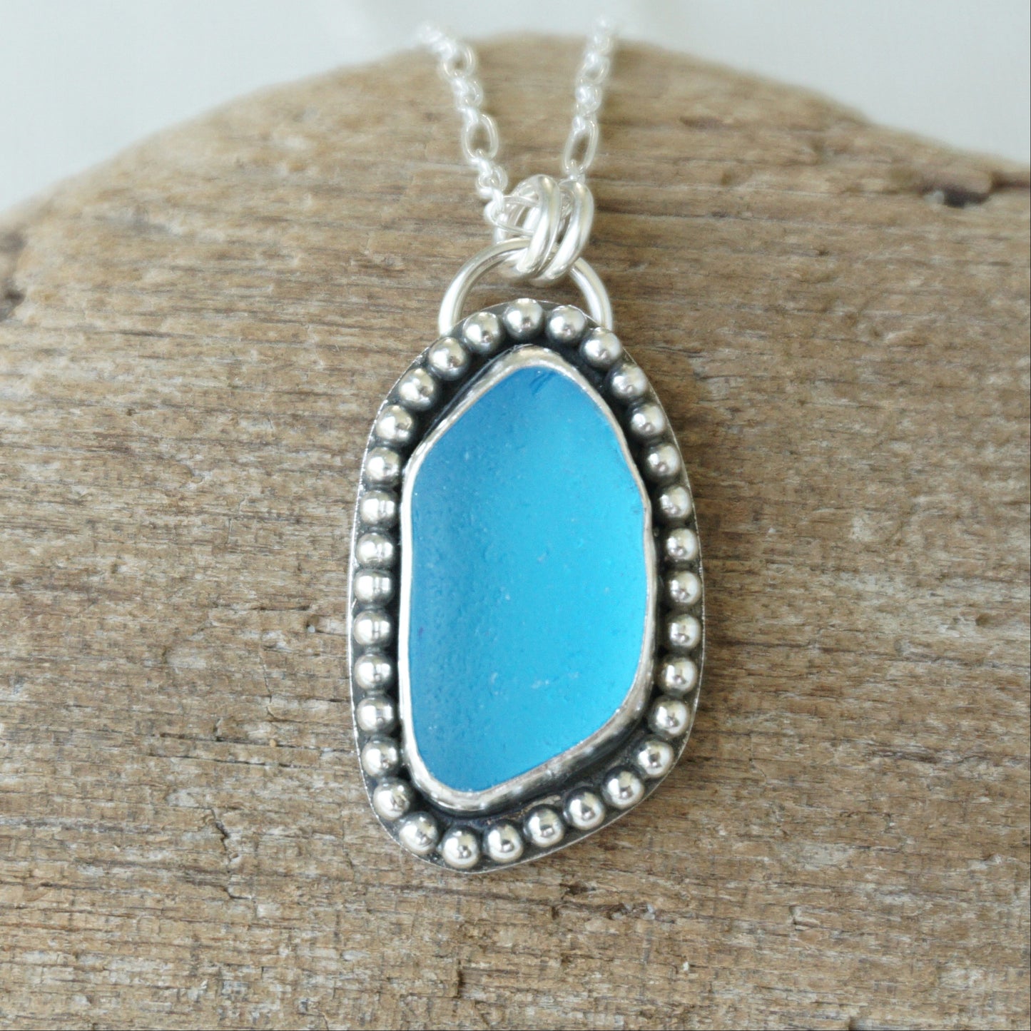 A silver-toned sea glass pendant necklace with bright turquoise blue sea glass, displayed on a textured piece of wood.