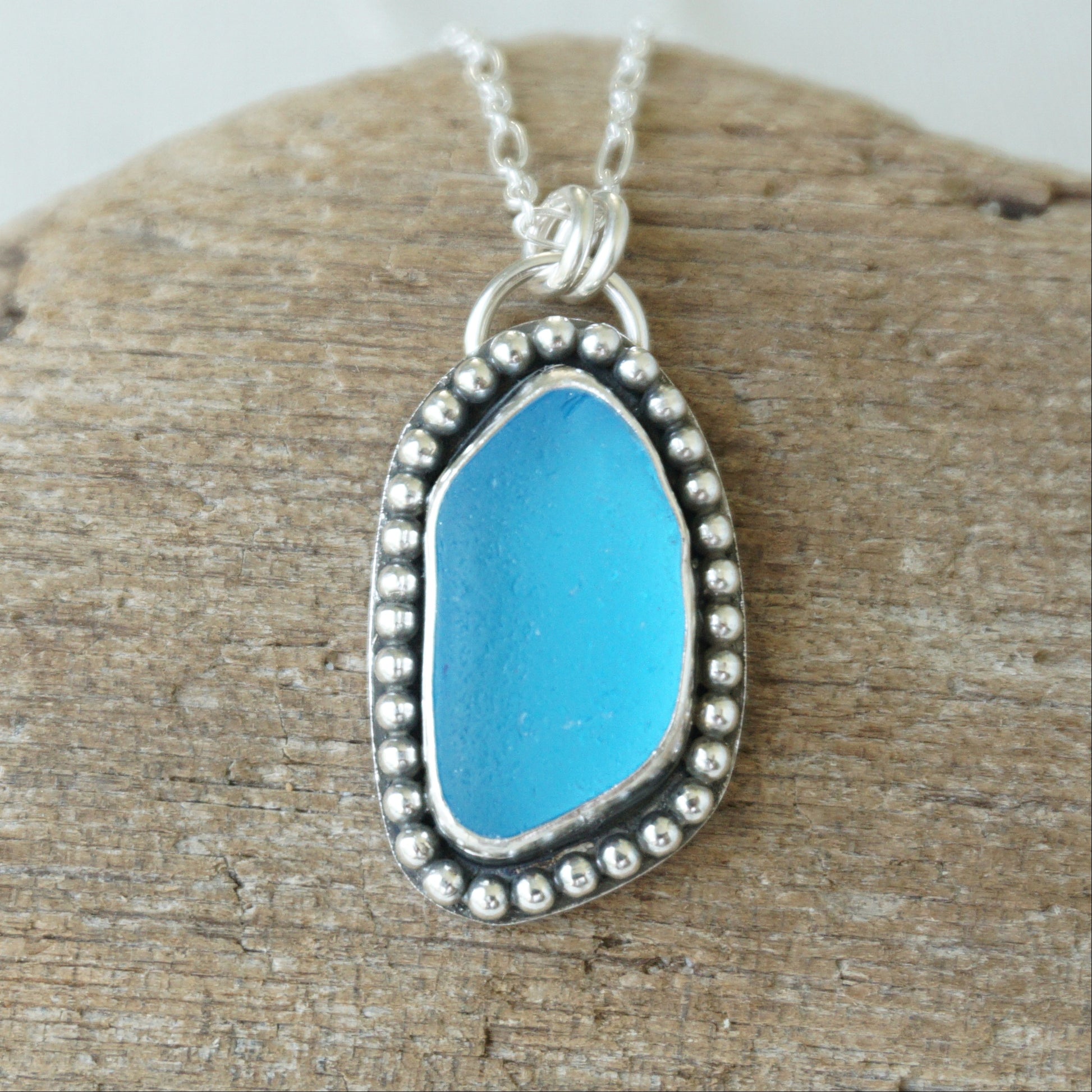 A silver-toned sea glass pendant necklace with bright turquoise blue sea glass, displayed on a textured piece of wood.