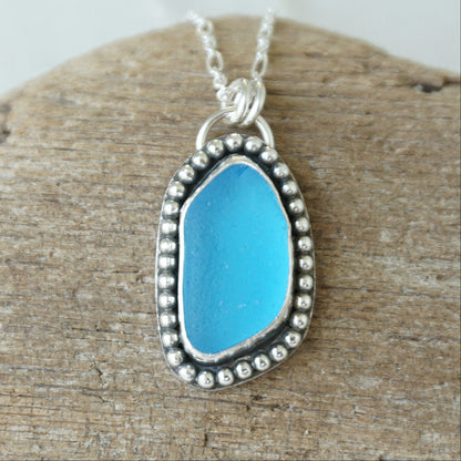 A silver-toned sea glass pendant necklace with bright turquoise blue sea glass, displayed on a textured piece of wood.