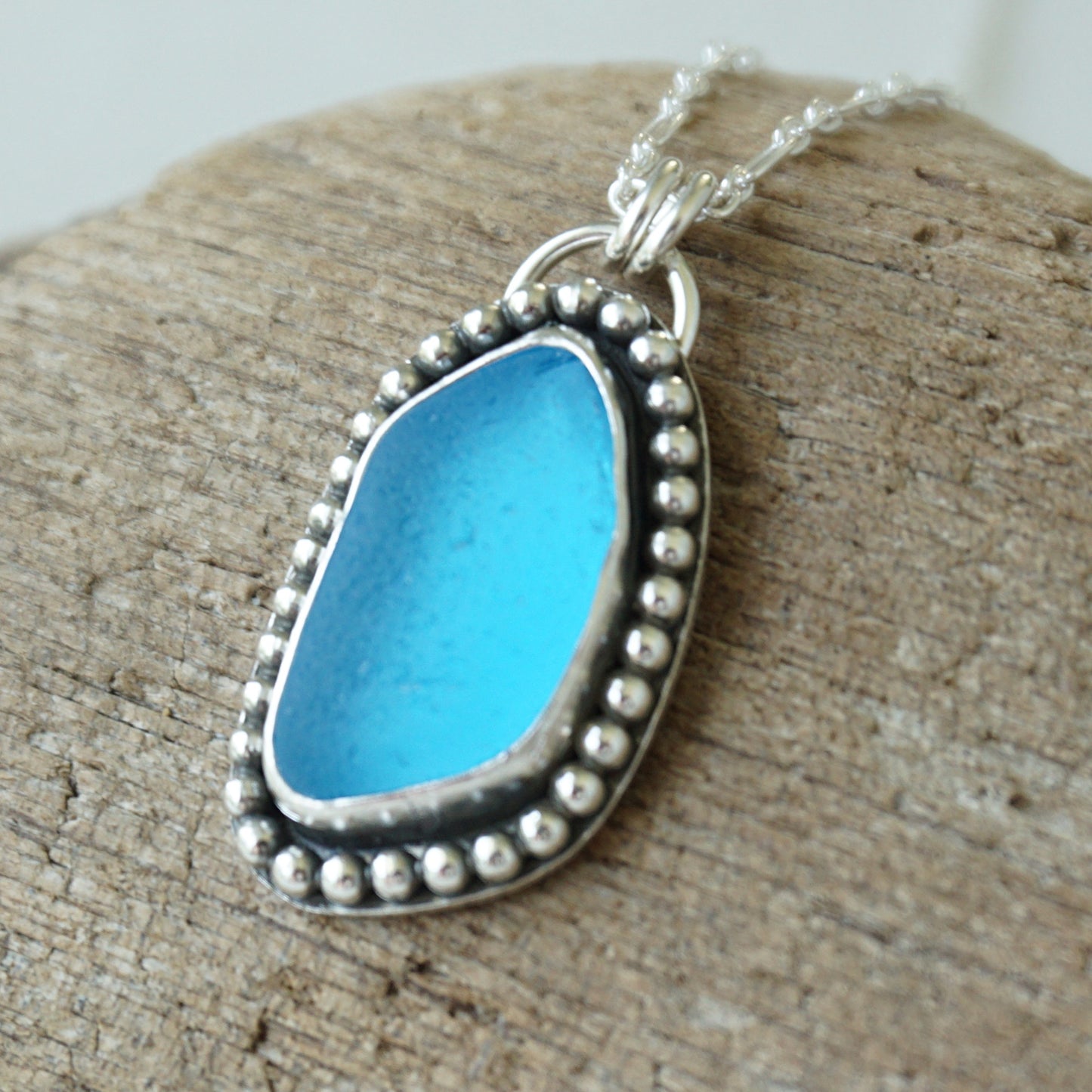 A sea glass pendant necklace featuring bright turquoise blue sea glass set in a silver beaded frame, resting on a textured piece of wood.