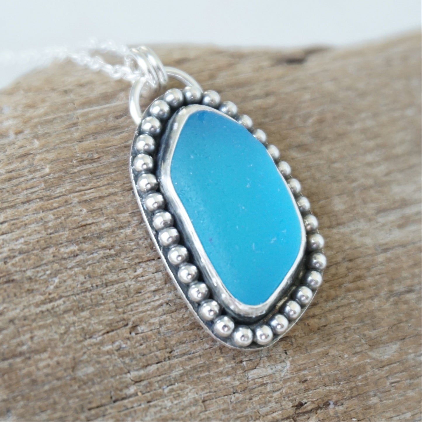 A silver-toned oval pendant necklace with bright turquoise blue sea glass, set in a beaded border and resting on a piece of weathered wood.