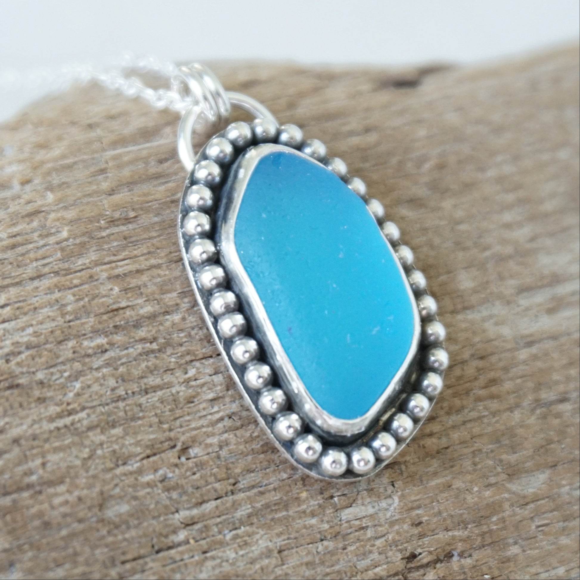 A silver-toned oval pendant necklace with bright turquoise blue sea glass, set in a beaded border and resting on a piece of weathered wood.