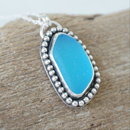 A silver-toned oval pendant necklace with bright turquoise blue sea glass, set in a beaded border and resting on a piece of weathered wood.