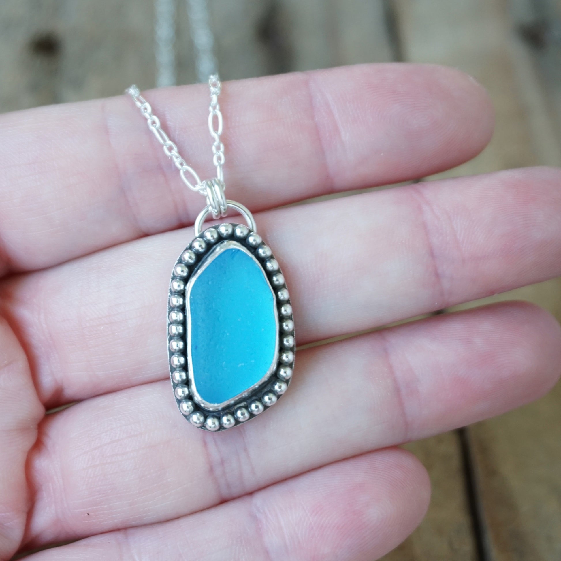A silver necklace with a turquoise blue sea glass pendant, held in a person's hand against a blurred wooden background.