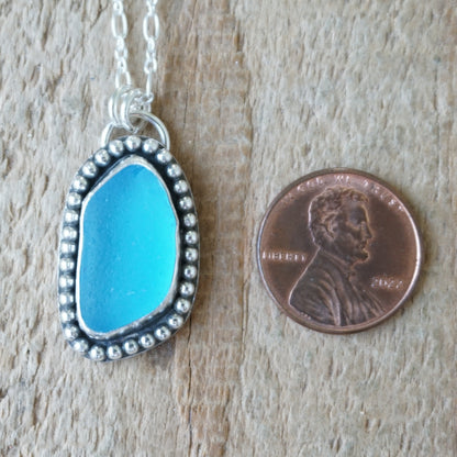 A silver-toned necklace with a turquoise blue sea glass pendant, shown next to a US penny for scale on a light wood background.