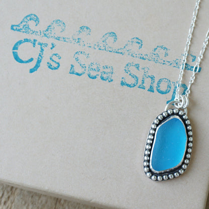 A silver-toned necklace with a teardrop-shaped bright blue glass pendant, displayed on a gift box