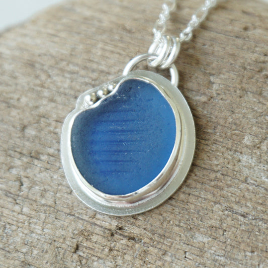 A round silver pendant necklace featuring a textured blue sea glass center, resting on a rustic, weathered wood surface.