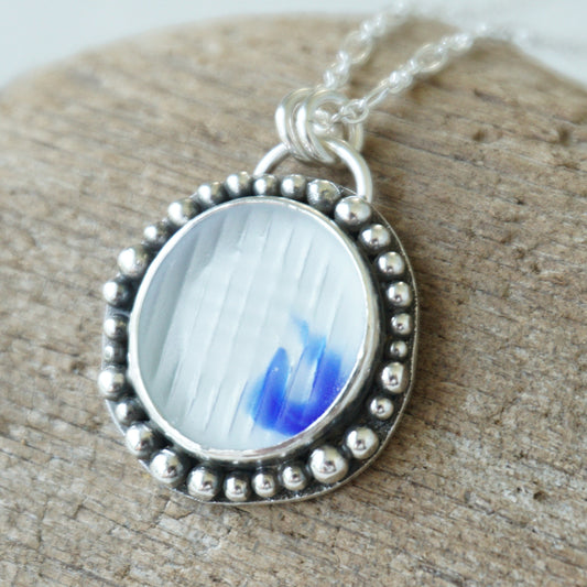 A silver pendant necklace with a beaded border and a clear and cobalt blue sea glass, displayed on a textured wood surface.