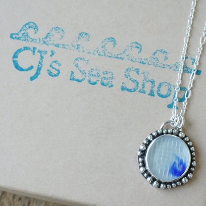 A silver pendant necklace with a beaded border and a clear and cobalt blue sea glass, displayed on a gift box