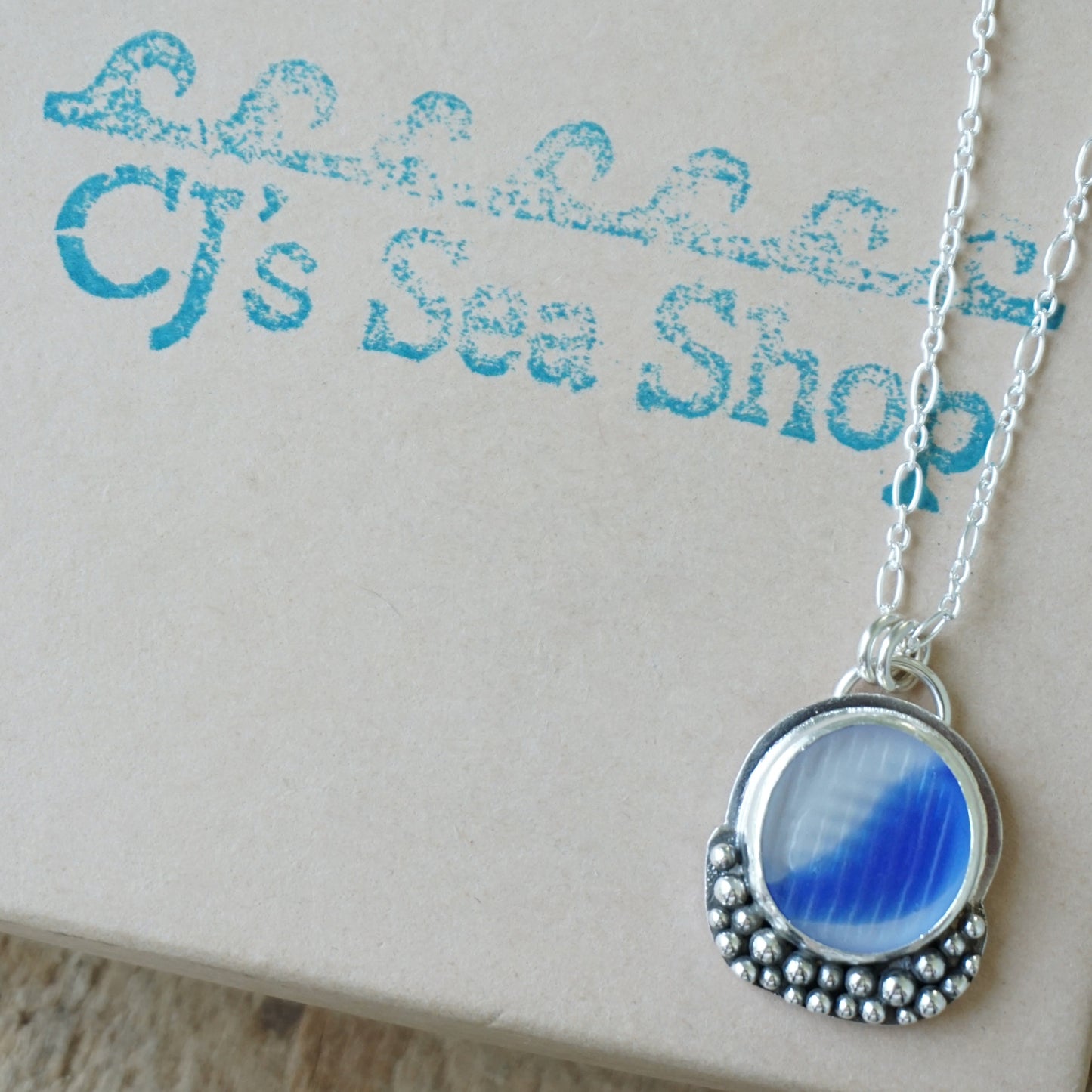 A silver pendant necklace with a round blue and clear marbled sea glass centerpiece, displayed on a gift box