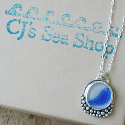 A silver pendant necklace with a round blue and clear marbled sea glass centerpiece, displayed on a gift box