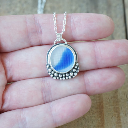 A silver pendant necklace with blue and clear marbled sea glass, held in a person's hand against a wooden background.