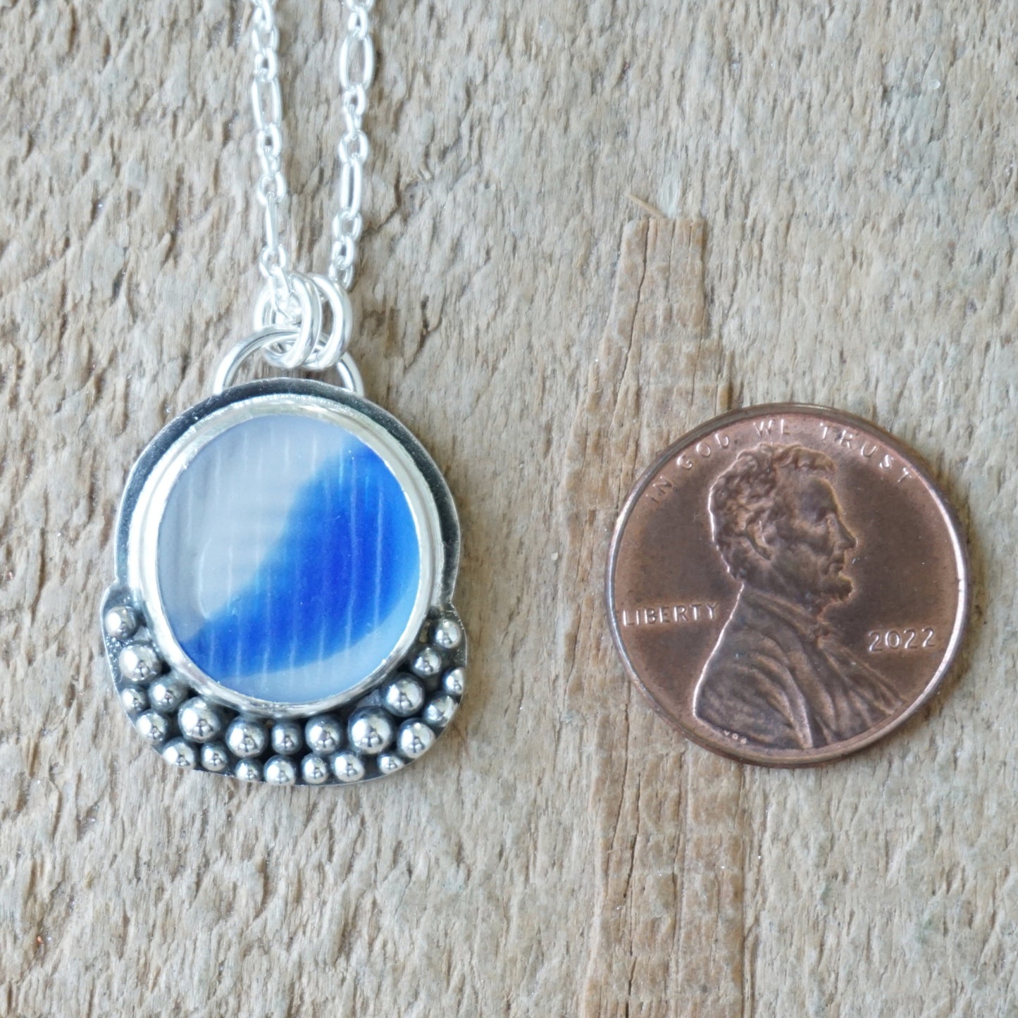 A silver pendant necklace with a round blue-and-clear marbled sea glass piece, shown next to a U.S. penny for scale