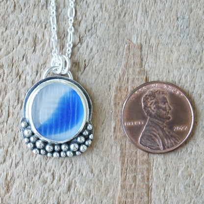A silver pendant necklace with a round blue-and-clear marbled sea glass piece, shown next to a U.S. penny for scale