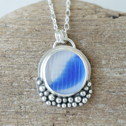 A silver pendant necklace with a round blue and clear marbled sea glass centerpiece, resting on a textured wooden surface.