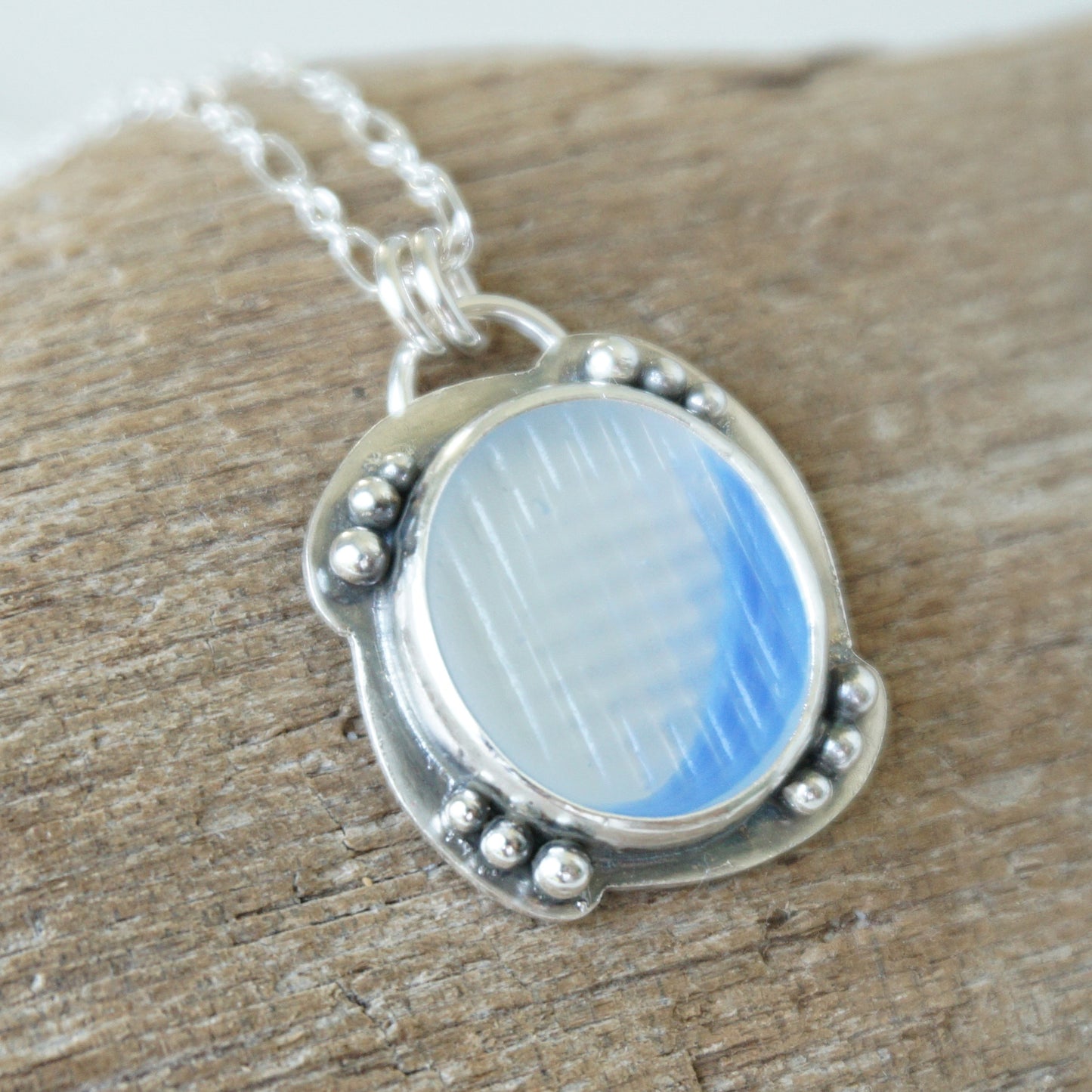 A silver pendant necklace with a round blue and clear marbled sea glass centerpiece, resting on a textured wooden surface.