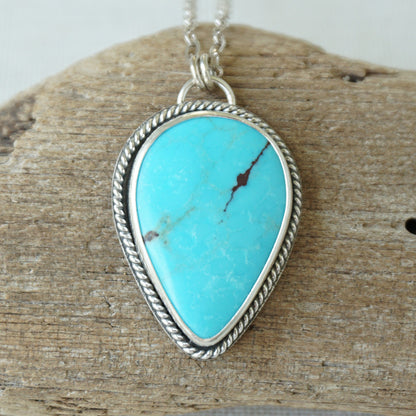 Sterling silver pendant with teardrop shaped turquoise stone, against a wooden background