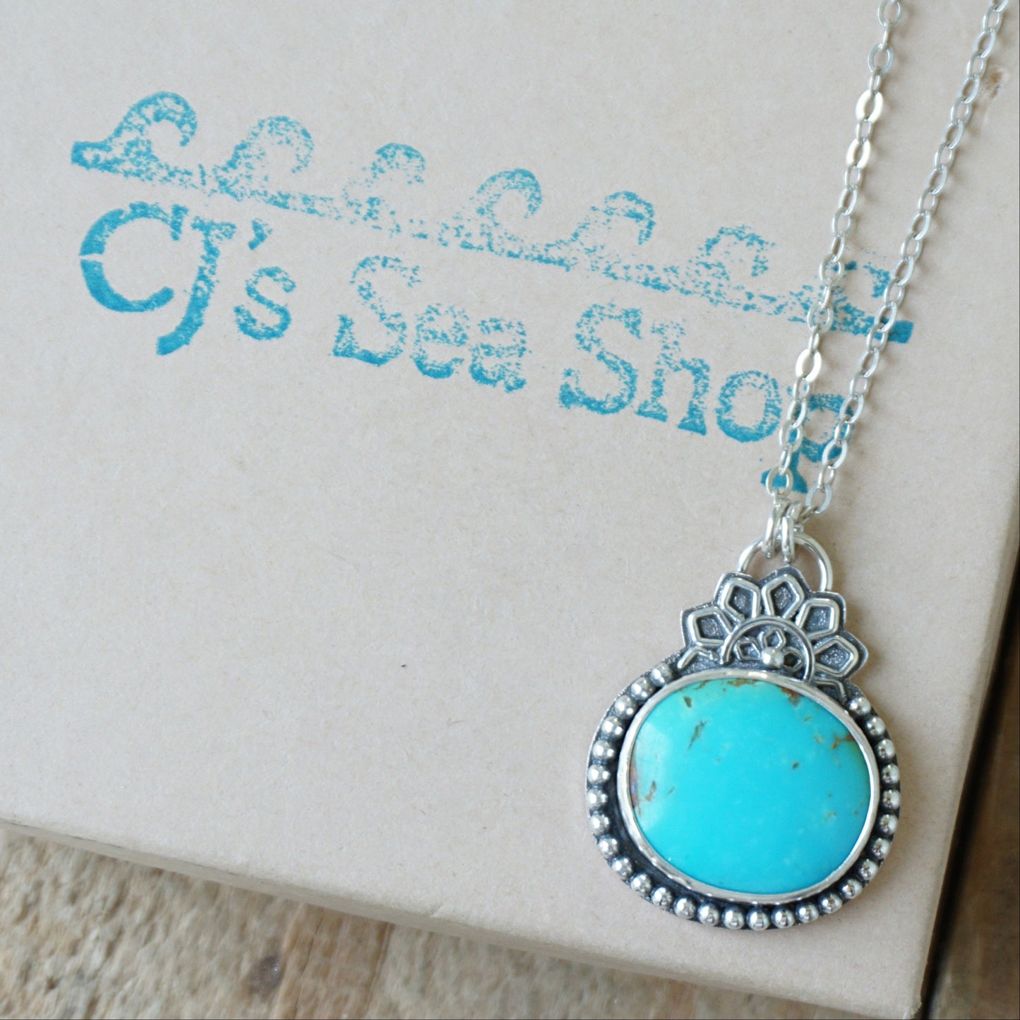A turquoise pendant set in sterling silver with silver floral accents, displayed on a gift box