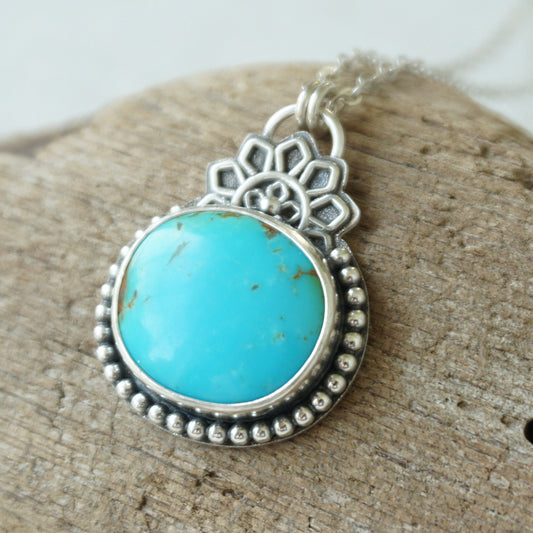 A turquoise pendant set in sterling silver with silver floral accents, against a wooden background