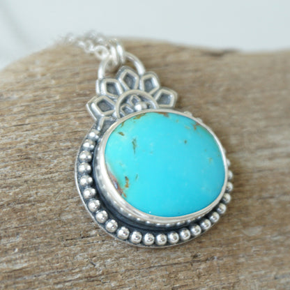 A turquoise pendant set in sterling silver with silver floral accents, against a wooden background