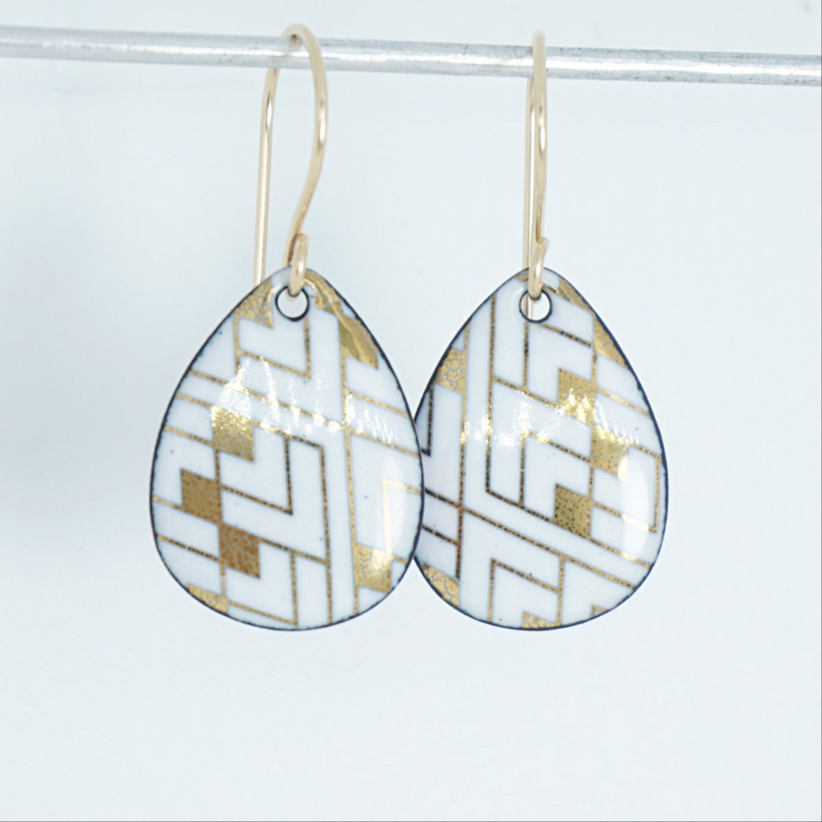 A pair of teardrop-shaped drop earrings with a white and gold geometric pattern, hanging from gold fishhook wires against a white background.