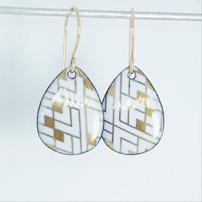 A pair of teardrop-shaped drop earrings with a white and gold geometric pattern, hanging from gold fishhook wires against a white background.