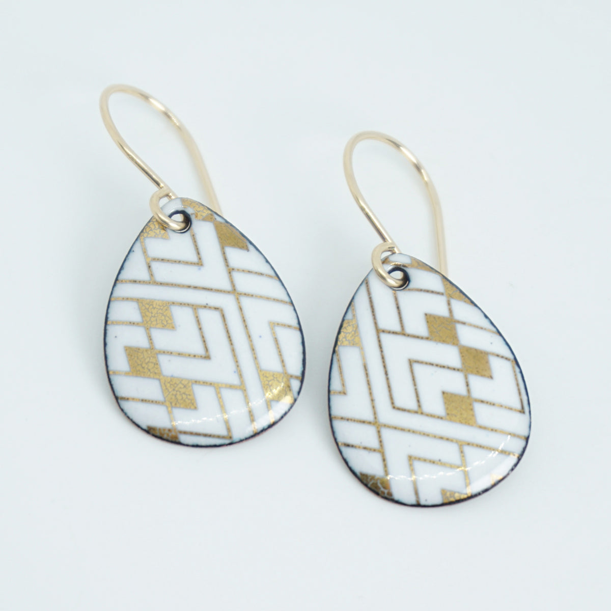 A pair of teardrop-shaped drop earrings with a white and gold geometric pattern, suspended from gold fishhook wires on a white background.