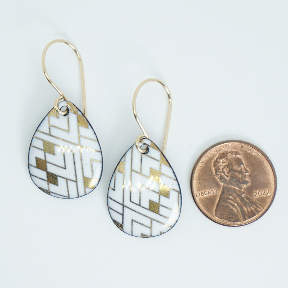 A pair of teardrop-shaped white enamel pendants featuring a gold geometric pattern, shown next to a US penny for scale on a white background.