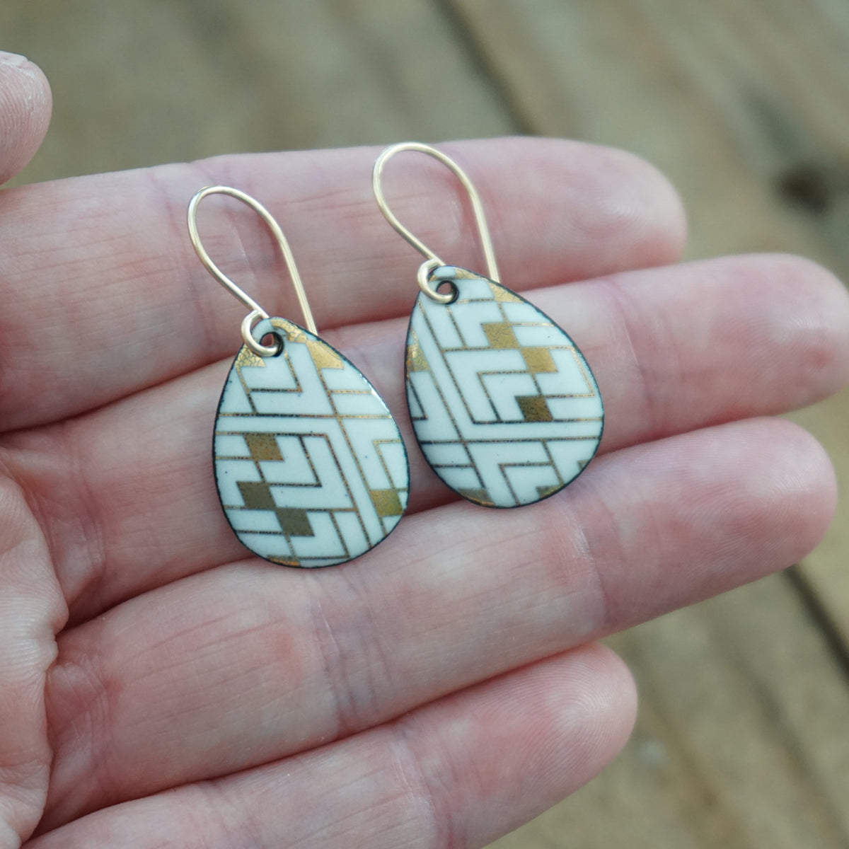 Hand holding a pair of teardrop shaped white enamel earrings with gold geometric pattern, against a wooden background