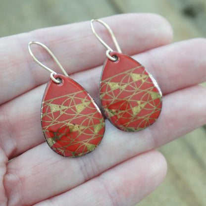 A pair of teardrop-shaped drop earrings with a red and gold geometric pattern, held in a person's hand. The earrings feature gold fishhook ear wires.