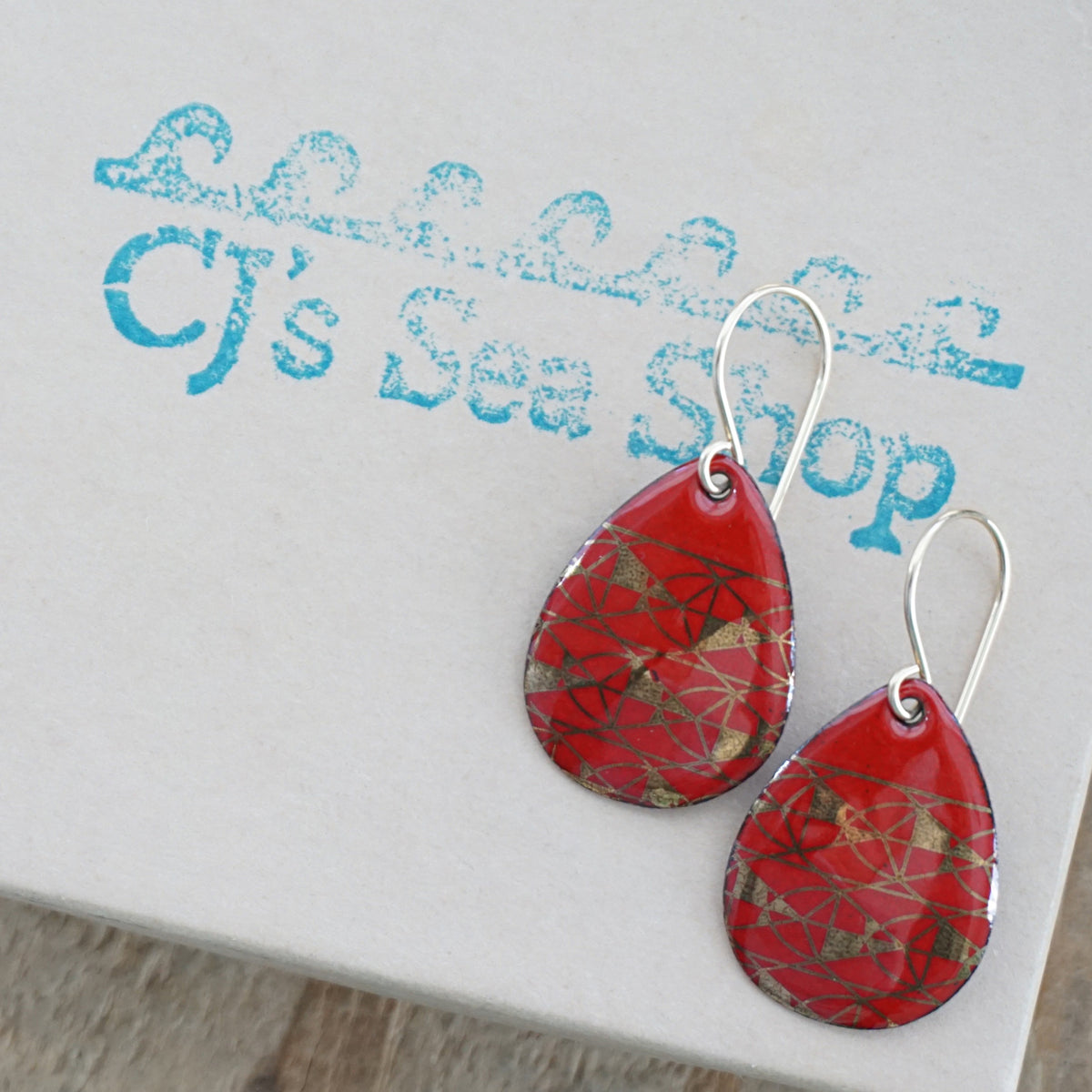 Teardrop shaped, red enamel earrings with gold geometric designs, displayed on a gift box
