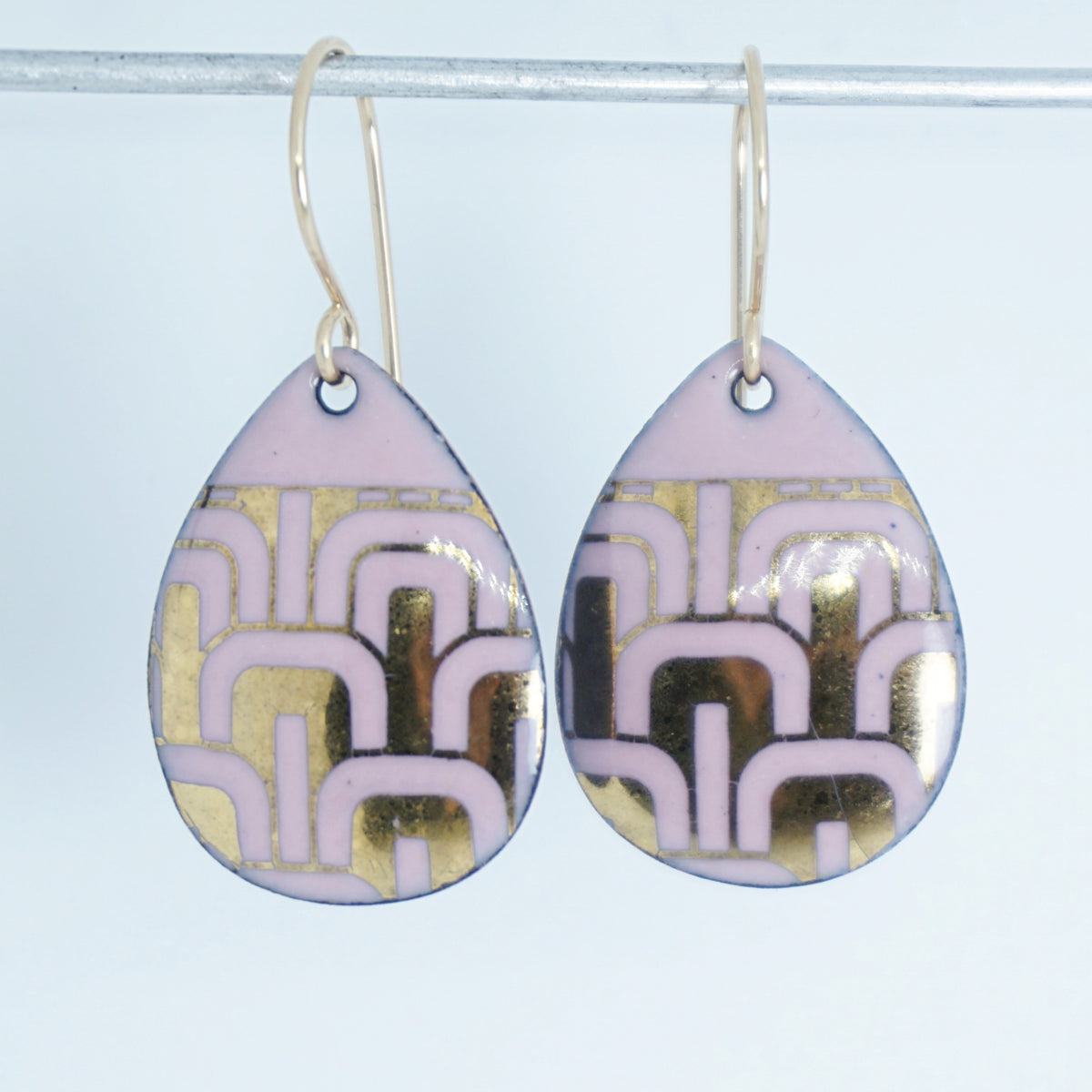 A pair of teardrop-shaped drop earrings with a light pink and gold geometric pattern, hanging from gold-tone hooks against a white background.