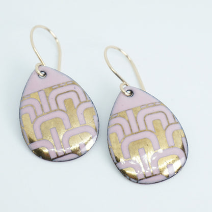 A pair of teardrop-shaped drop earrings with a light pink and gold geometric pattern, suspended from gold-tone wire hooks against a white background.
