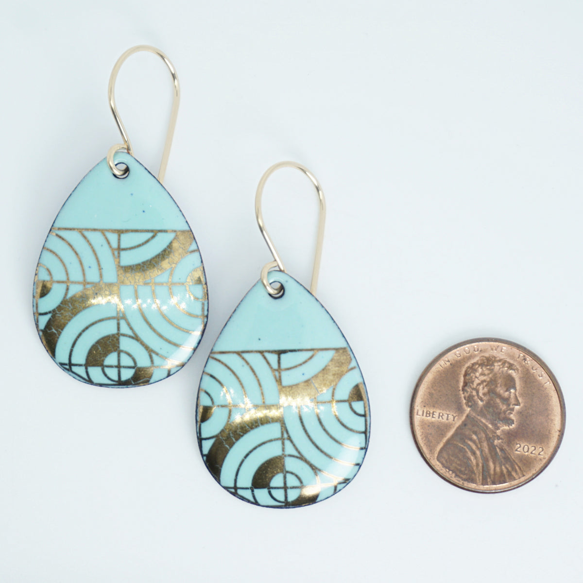 Teardrop shaped, seafoam green enamel earrings with gold geometric designs, next to a penny for scale, against a white background